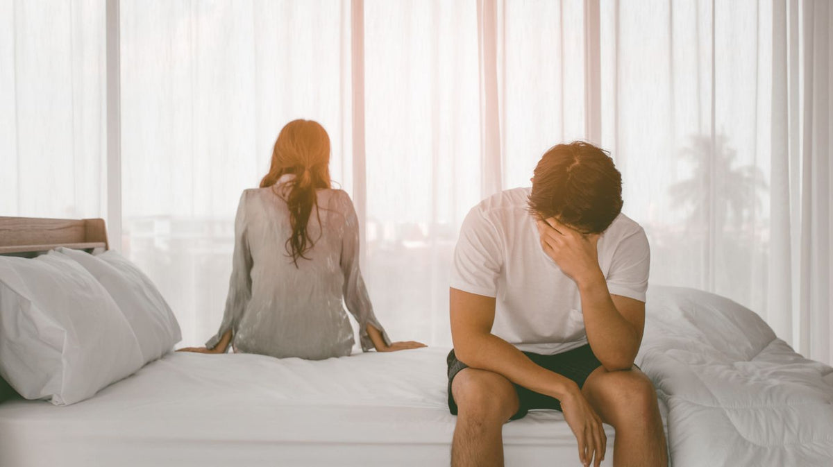A man in a white t-shirt and dark shorts sits dejectedly on a white bed, while a woman in a sheer gray top sits on the other side of the bed, Conflict in relationship